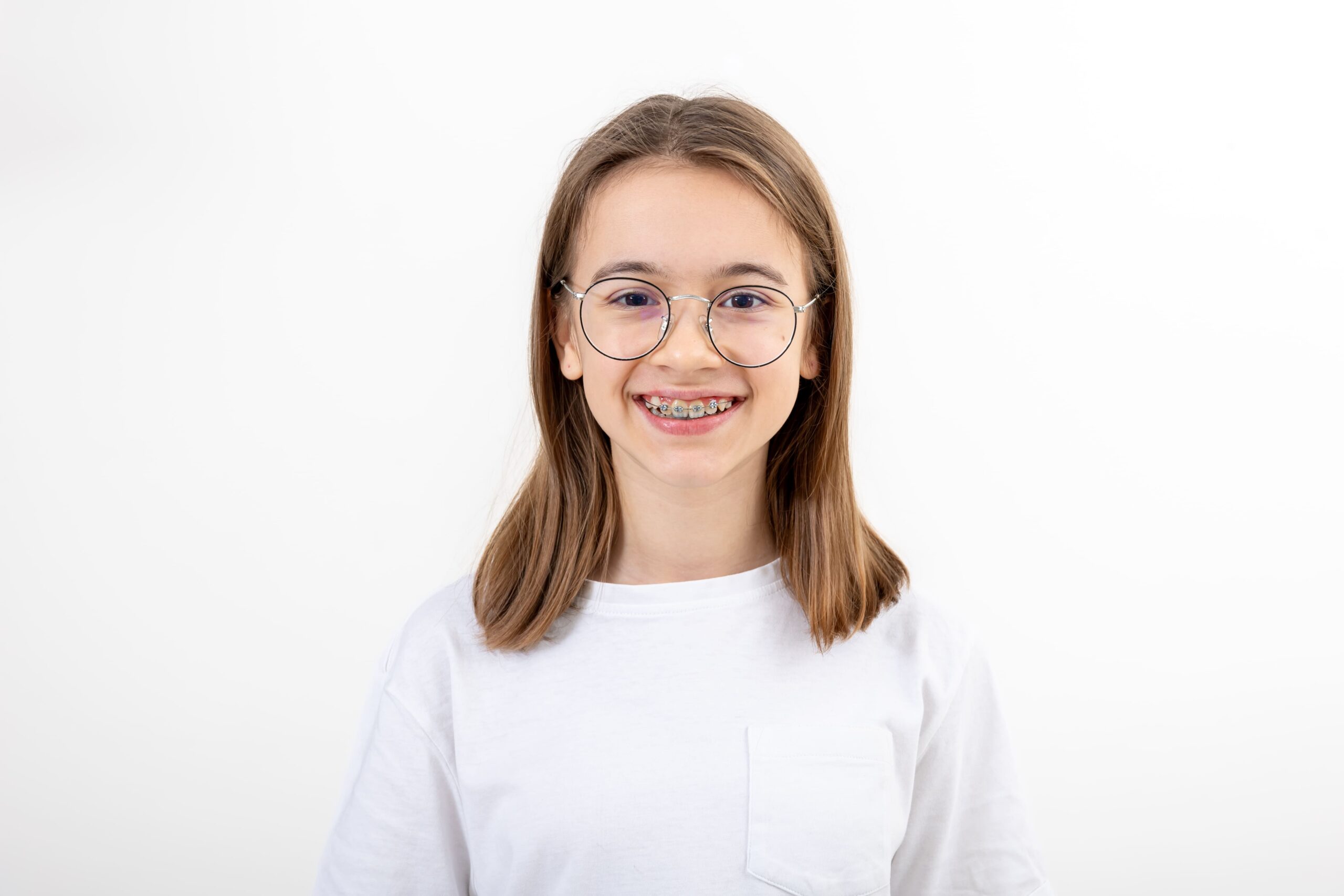 A young person with long brown hair, glasses, and braces smiles at the camera while wearing a white t-shirt against a plain white background.